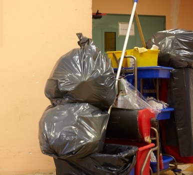 The image displays a collection of black rubbish bags piled on top of each other, resting on a red trolley trolley with a metal frame, located indoors against a yellow wall. The bags appear to be filled with waste material and are of standard size, with some leaning to the sides and showing creases on the plastic surface. Behind the bags, there is a blue plastic bin with a yellow shovel or broom handle resting on it, positioned vertically, and partially obscured by black trash bags. To the right, additional black rubbish bags are visible, stacked against a wall or larger container. The environment suggests a storage or disposal area, with the waste material likely accumulated for collection or removal as part of commercial waste management services in Feltham, nearby London. The lighting is neutral, indicating indoor artificial illumination, and the scene captures a typical rubbish collection setup involving waste bags and equipment for professional rubbish removal, as used by companies like Commercial Waste Feltham.
