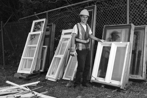 A young male worker wearing a safety helmet and reflective vest stands outdoors on a grassy area near a chain-link fence, surrounded by various white window frames with glass panes leaning against the fence and placed on wooden pallets. The window frames appear to be made of wood or UPVC, with some showing protective film or finish, and are arranged in an orderly manner. The ground is scattered with additional smaller frame parts and wooden debris, indicating a site for window disposal or replacement. The setting suggests a local waste removal operation, typical of services offered by Commercial Waste Feltham, with natural daylight illuminating the scene. The background includes trees and a clear sky, providing a natural context for this exterior rubbish collection scene in the outskirts of Feltham, near London.