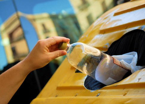 A person's hand is shown holding a transparent plastic bottle filled with clear liquid, about to dispose of it into the large yellow rear-opening hatch of a waste collection vehicle or bin, which has a circular opening in the centre filled with crumpled paper and other waste materials. The background features an urban environment with blurred buildings and a clear sky, indicating the waste is being collected or managed in a town area. The scene emphasizes the process of rubbish disposal, with the plastic bottle as the primary object, along with the textured, shiny surface of the bottle and the yellow painted exterior of the waste vehicle or container, consistent with local rubbish removal services such as those offered in Feltham, London postal district TW13. The environment suggests an outdoor urban setting during daytime, with natural lighting illuminating the scene's details, relevant to commercial waste collection activities conducted by companies like Commercial Waste Feltham.