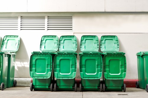 A row of ten green wheelie bins with closed lids, positioned against a white building wall with horizontal vent slats near the top. The bins are uniform in size and have black wheels at the base. They are set on a grey concrete pavement, with a red-painted curb visible behind the bins. The scene is outdoors, likely in a commercial or residential area in Feltham, with natural daylight illuminating the bins and surrounding environment. The image appears to relate to waste collection or rubbish management services provided by Commercial Waste Feltham, targeting efficient rubbish disposal solutions in the local area.