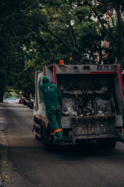 A waste collection worker dressed in green protective clothing and a helmet is standing on the pavement beside a large, industrial rubbish collection truck with an open rear compartment, which is visi