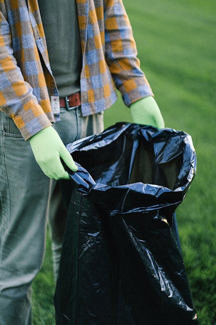 A waste collection worker dressed in green protective clothing and a helmet is standing on the pavement beside a large, industrial rubbish collection truck with an open rear compartment, which is visi